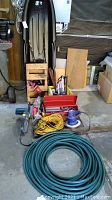 Photo showing black plastic sled standing vertically in corner, fishing poles in yellow crates, wooden box of tools, and coils of green garden hose and yellow extension cord on floor near a power sander and toolbox.
