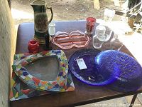 Photo of the lot showing the two blue platters, pink divided plate, cranberry glass vase with chip, ceramic pitcher, small red glass vase, and various clear glass pieces on a wooden table.