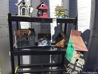 Photo of seven various bird houses on black metal shelving unit outdoors in daylight, showing diverse architectural styles and materials.