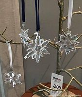 Four Swarovski crystal Christmas ornaments displayed on a decorative branch, showing varying snowflake and star shapes with ribbons for hanging.