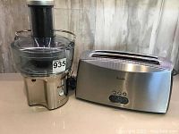 Juicer and toaster side by side on table, showing juicer's body with black top chute and clear juice container and the toaster with wide slot and brushed stainless steel finish.