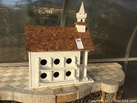 Front view of white wooden bird house with six round openings and brown shingle roof with small steeple.