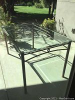 Rectangular glass top table with black metal frame and clear glass top with shadows on outdoor concrete surface