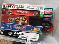 Photo showing an assortment of boxed board games: Sorry!, Monopoly electronic banking edition, Deluxe Edition Yahtzee, classic Monopoly, and Othello. Also includes 6 decks of kids playing cards and an electronic handheld game device.