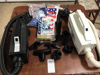 Black Oreck BB-280D vacuum cleaner, vacuum hose, multiple attachments including brushes, nozzles, connectors, and 2 unopened packages of vacuum bags on a wooden table.