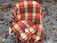 Full view of the folded vintage Woolrich blanket laid over a chair outdoors with light foliage and gravel on the ground.