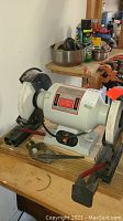 8 inch slow-speed bench grinder on a wooden workbench with grinding wheels and tool rests visible. Some miscellaneous tools nearby. Unit appears heavy and condition untested.