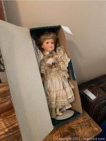 Porcelain doll in original box, showing full figure dressed in detailed white lace dress with colorful floral accents.