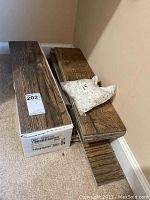 Photo of flooring tiles stacked on the floor next to a wall, with a white bag of spacers on top and a box labeled with lot number 202.