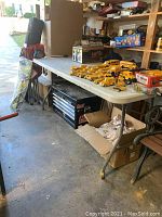 View of the folding table showing white plastic top with several small scuffs and dust. Metal legs and frame visible, set up in garage with other miscellaneous items around but not included.