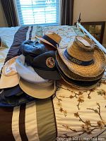 Pile of assorted hats including straw cowboy hats with decorative bands, fabric baseball caps, and flat caps on a bed with a floral and striped comforter