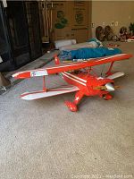 Side view showing the red biplane with white stripes and blue accents on tail, wooden propeller, and landing gear with wheels.