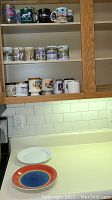 Photo of 11 assorted mugs displayed on two shelves inside a wooden cabinet above a kitchen counter, along with two plates placed on the counter.