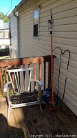 Outdoor porch view showing metal chair, shepherd hooks, tree pruner, propane torches and bird feeders arranged against house wall.