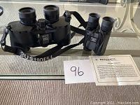 Photo showing the two pairs of black Bushnell binoculars side by side on a glass table, accompanied by the warranty sheet and a number card labeled 96.