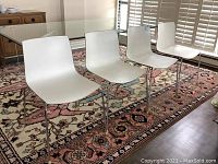 Four white chairs in a row on a patterned rug, showing front view of plastic seats and chrome legs