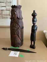Photo showing hinged wine bottle holder carved as a crowned woman, thin African-style wooden female figure, and a metal letter opener on a flat surface.