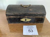 Front view of the closed small antique jewelry box showing black worn exterior, brass keyhole plate and handle.