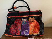 Front view of the tote bag showing colorful cat artwork in red, purple, and orange cat figures with patterns on black fabric background, and handles visible.