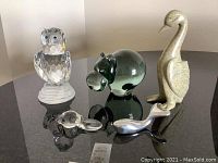 Five animal figurines on a black reflective surface: crystal owl, greenish glass hippopotamus, two small glass birds, and tall soapstone bird.