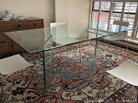Wide view of entire clear glass rectangular dining table on patterned rug with white chairs around it.