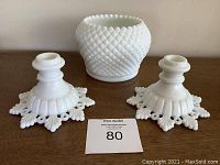 Two white milk glass candlesticks with ornate star-like bases next to a round milk glass bowl with hobnail pattern exterior, all placed on a brown surface.