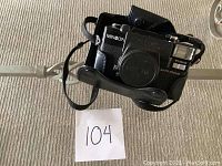 Front view of Minolta Hi-Matic AFD camera showing lens with cap and attached black carrying case on carpet surface.