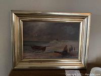 Framed oil painting showing coastal scene with boat and two figures near shore, with broad gold frame.
