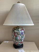 Full view of the lamp with beige pleated shade and floral ceramic base on wooden stand.