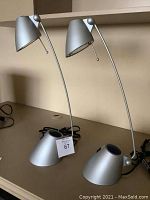 Pair of silver metal desk lamps with curved adjustable necks and cone-shaped heads on a shelf.