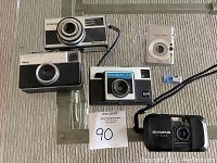 Top view of all five vintage cameras on a carpeted surface: Zeiss Ikon Ikromatic A, Kodak Instamatic X15, Kodak Instamatic 133, Olympus 35 SP III, Canon PowerShot SD1100 IS