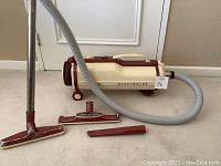 Side view of vintage Electrolux cylinder vacuum with hose and three attachments on floor.