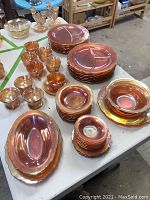 Photo showing overall collection of carnival glass dinnerware including various sized bowls, divided plates, small plates, and cups all with iridescent amber finish.
