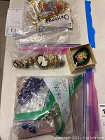Photo showing multiple zip-lock bags containing assorted holiday-themed costume jewelry, cameo pieces, and sets of beads and earrings. A hand-painted floral brooch in a box is also visible.