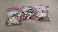 Three ziplock bags of costume jewelry necklaces on a concrete surface. Bags are labeled as necklaces.