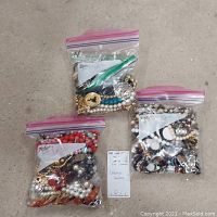 Three plastic bags containing assorted costume necklaces and bracelets on concrete floor.