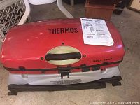 Red Thermos grill with latch, logo, and instruction manual on top.