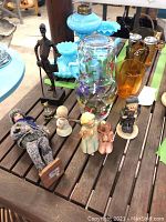 Shows the overall lot of various figurines including fabric doll, metal man figure, two Hummels, and glassware