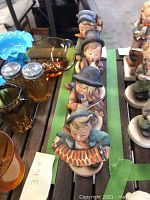 Photo showing four different Hummel figurines arranged in a row, displayed on a wooden surface with some glassware in the background.