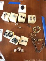 Multiple pieces of jewelry displayed on beige boxes and wood table. Includes earrings in various styles, bracelets, and pins. Some earrings have black, turquoise, or pastel stones. Bracelets are silver-tone with link designs.