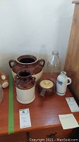 Photo of a collection of kitchen stoneware jugs, a large two-handled brown bean pot, a clear glass bottle, and a dark brown pottery vinegar cruet with a lid and spout.