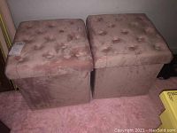 Two matching pink velvet-like upholstered storage ottomans with tufted tops. Square shape, cushioned tops with button tufting, set on a light pink carpet.