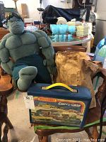 Large green plush Incredible Hulk doll seated next to vintage blue Matchbox Carry Case with yellow handle on wooden chair, background shows garage environment and papers.