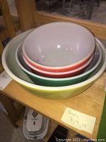 Four nested vintage Pyrex mixing bowls in yellow, green, and orange-red tonal colors with white interiors, placed on a wooden shelf.