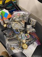 Photo of multiple assorted wristwatches in plastic bags and loose on a black surface showing variety of styles, bands, and materials