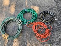 Four coiled hoses laid out on pavement: green garden hose, greenish-gray garden hose, black garden hose, and an orange extension cord.