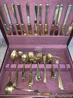 Full view of the open silverware chest showing gold tone forks, spoons, knives, and serving utensils placed in their slots with purple velvet lining.