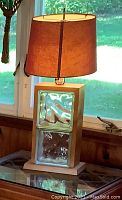 Full view of the lamp placed on a glass surface near window showing the wooden frame, two glass blocks, and brown fabric shade with light on.