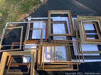 Group of 19 large art frames arranged outdoors, showing multiple gold and brown wooden frames without glass, including one metal frame and metal corners.