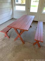 Full view of vintage wooden picnic table with two benches, painted dark reddish brown, set on a covered patio floor.